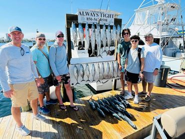 Group of six men showing off a big catch of fish on a dock under clear skies.
