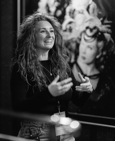 A smiling woman with curly red hair speaks animatedly in front of colorful artwork.