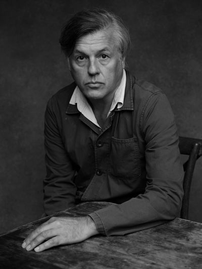 Mature man in blue jacket sitting thoughtfully at a rustic wooden table.