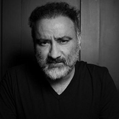 Serious man with gray beard and black shirt against dark background.