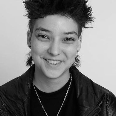 Smiling person with spiky hair wearing a black jacket and necklace.