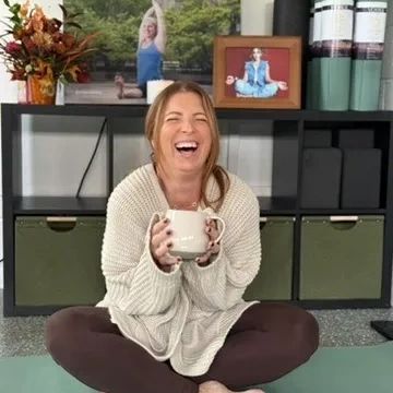A woman laughs joyfully sitting cross-legged on a yoga mat holding a mug.