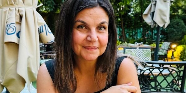 A woman smiling at an outdoor café with greenery in the background.