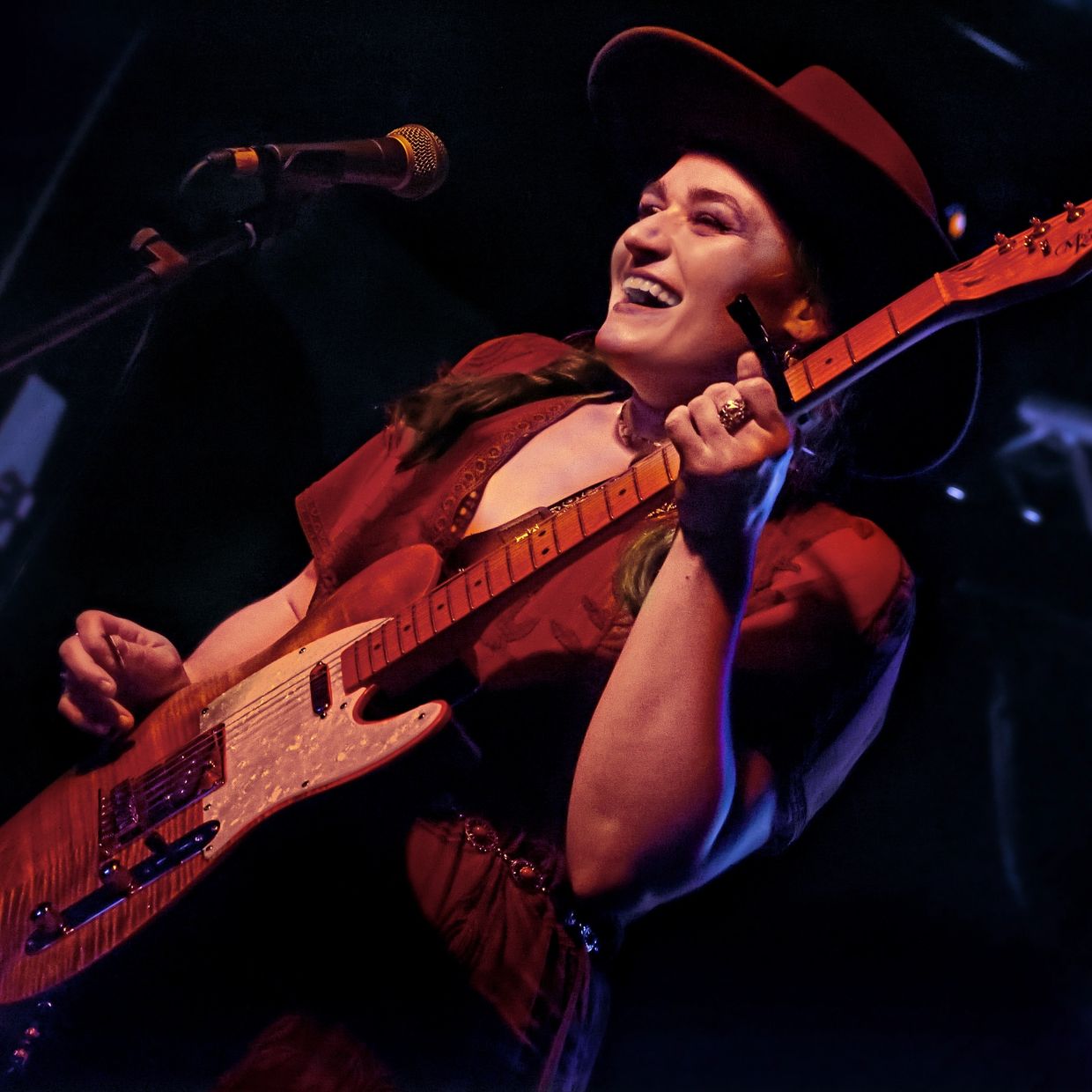 A joyful musician plays electric guitar on stage, wearing a hat and red outfit.