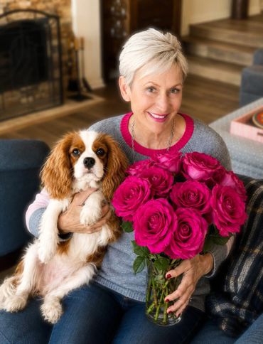 Smiling woman holding a dog and a bouquet of pink roses indoors.