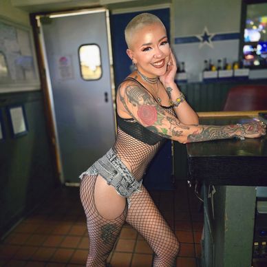Smiling woman with tattoos and fishnet outfit leaning on a bar counter.