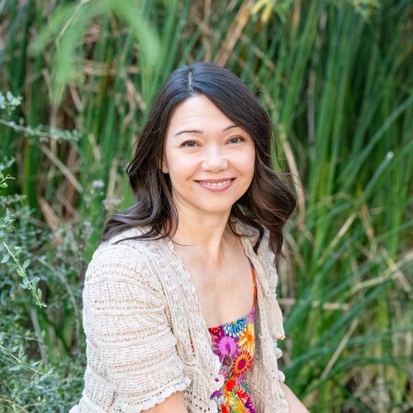 Smiling woman in a floral dress and beige cardigan outdoors with greenery.
