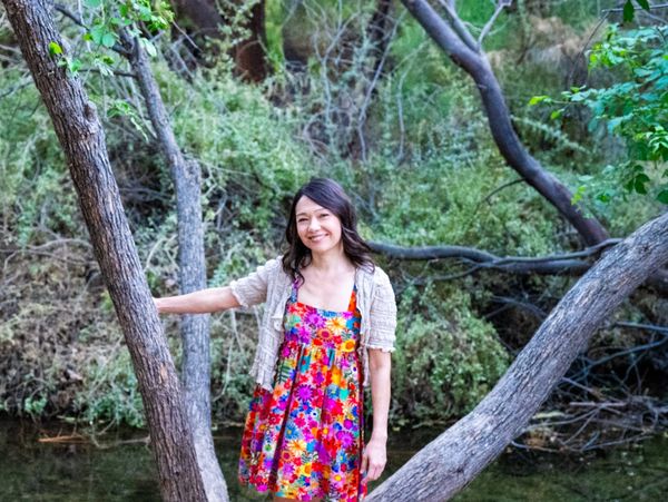 A woman in a colorful floral dress stands between tree branches in a forest.