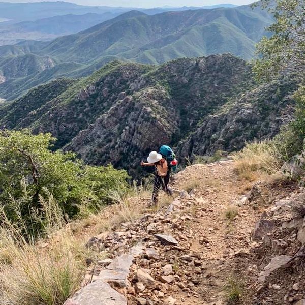 Hiker with backpack trekking a rocky mountain trail surrounded by green hills.