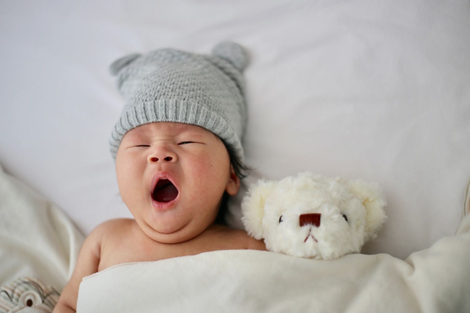 Yawning baby wearing a gray knit hat with bear ears, lying next to a white teddy bear.