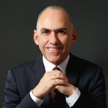 Smiling man in a dark suit with clasped hands.