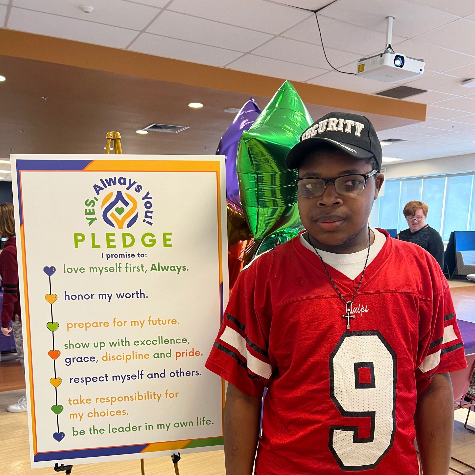 Man in red jersey standing beside a pledge sign indoors.