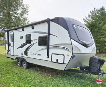 White Cougar travel trailer parked on grass with trees in the background.