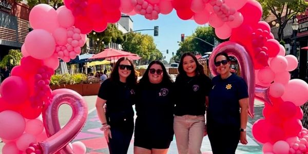 Four women standing under a pink balloon arch outdoors.