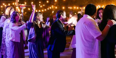 People dancing in a conga line at a festive outdoor party with string lights.