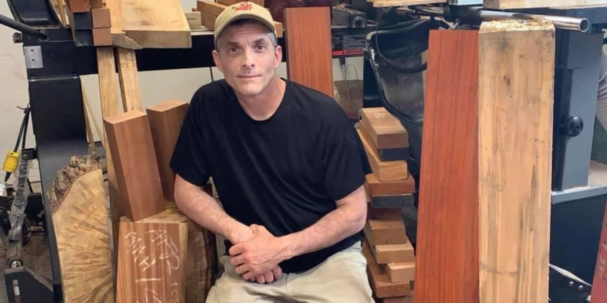 A man sits among various wooden planks and blocks in a workshop.