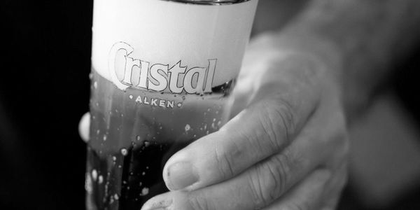 Hand holding a glass of Cristal Alken beer with foam on top.