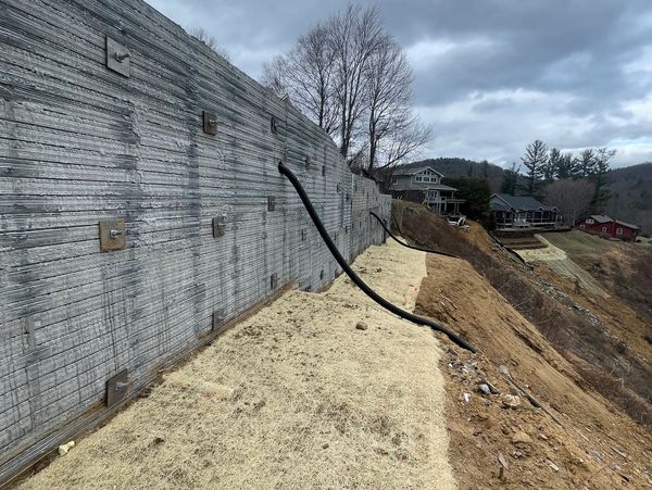 Concrete retaining wall with exposed pipes on a hillside under a cloudy sky.