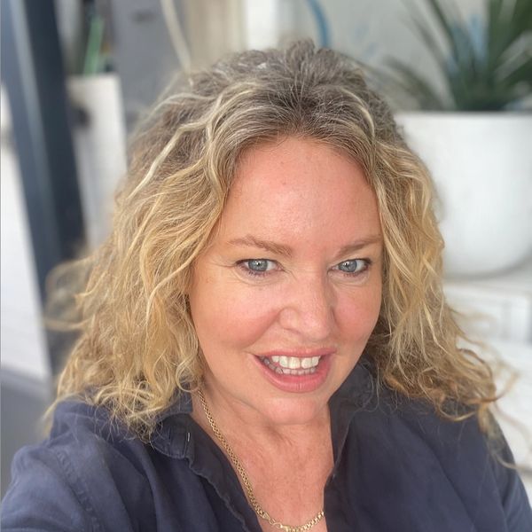 Smiling woman with curly blonde hair wearing a dark shirt and gold chain necklace.