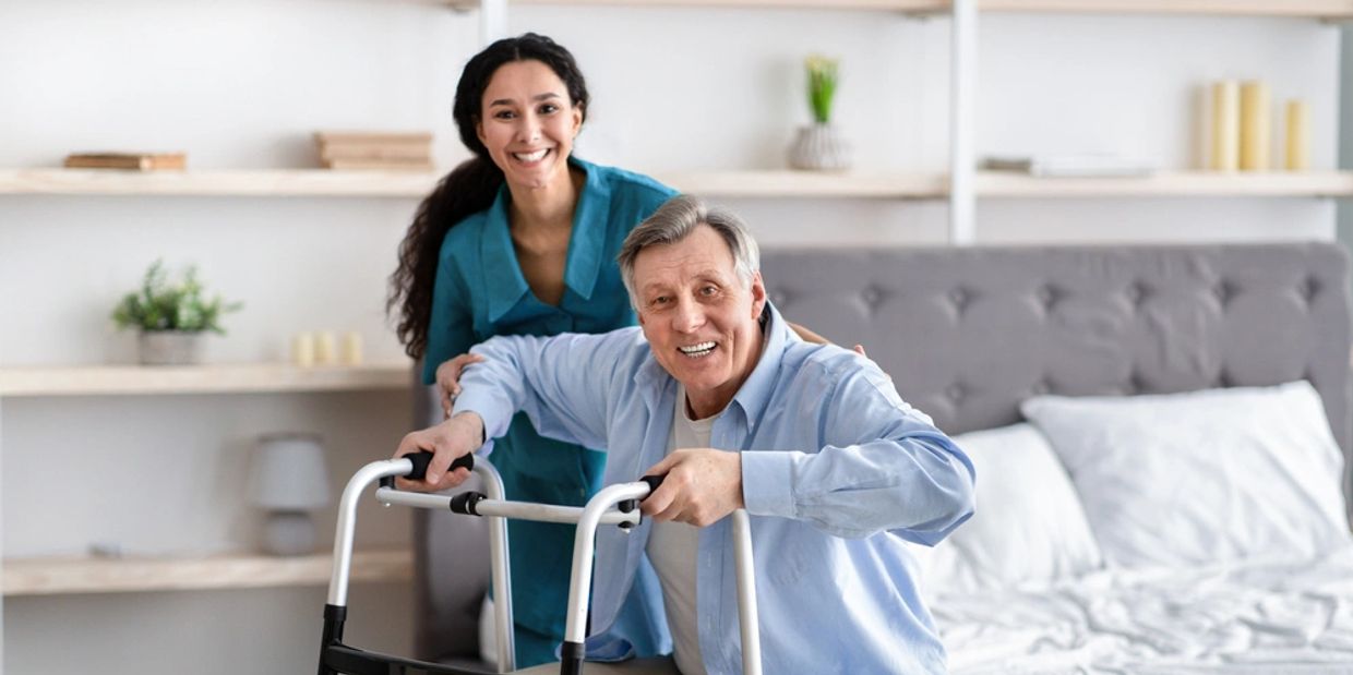 Female nurse helping elderly male with walking frame stand up from bed at home.