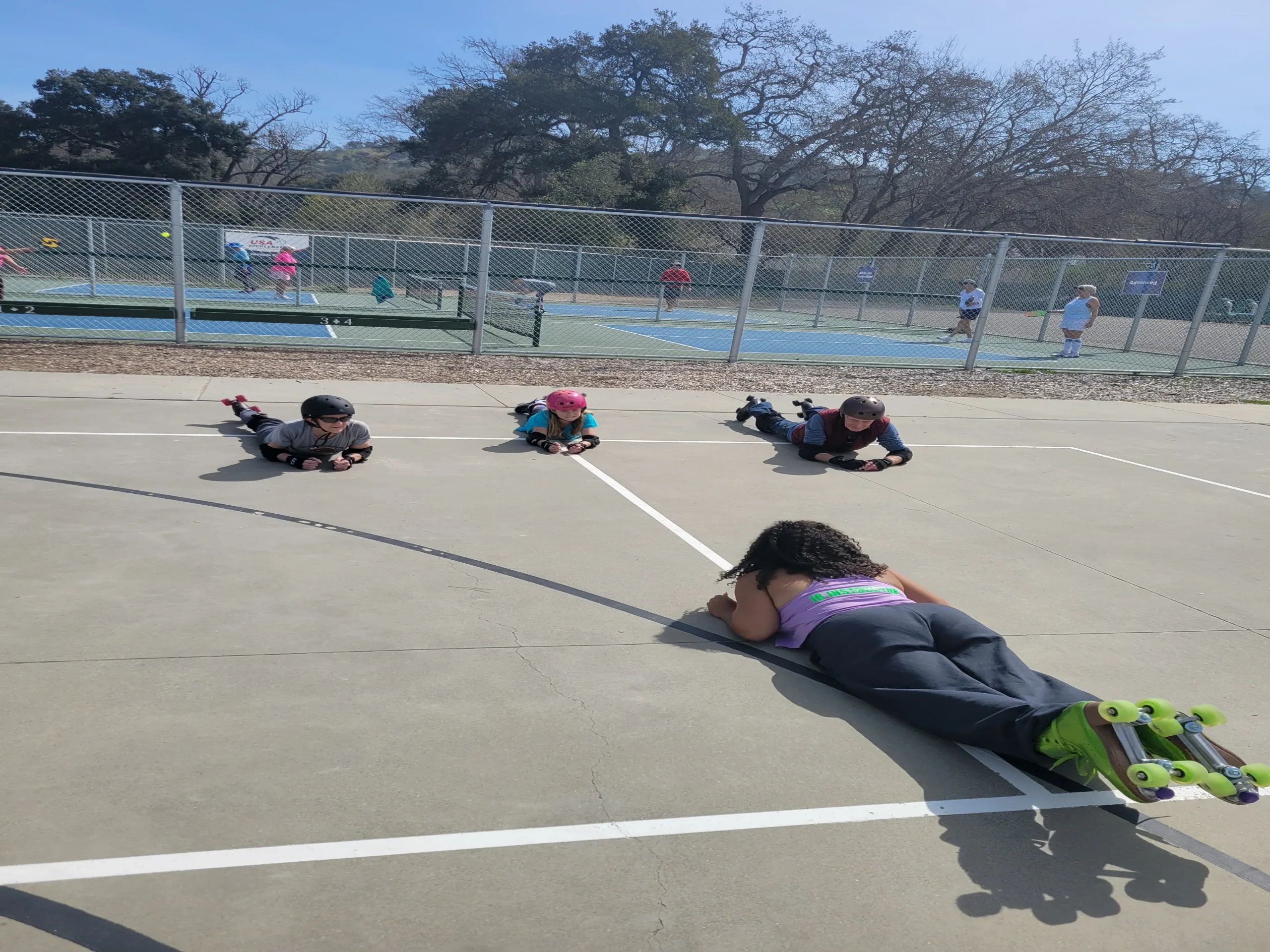 Three kids lying on the ground with helmets and roller skates, a coach in front.