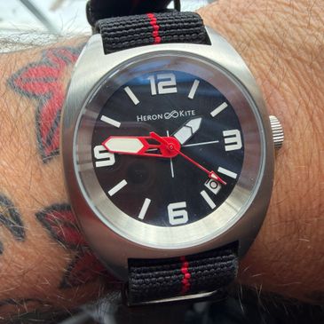 Wristwatch with a black dial and red-accented hands on a tattooed wrist.