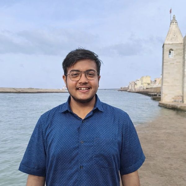 Young man in glasses smiling by the water near a historic tower.
