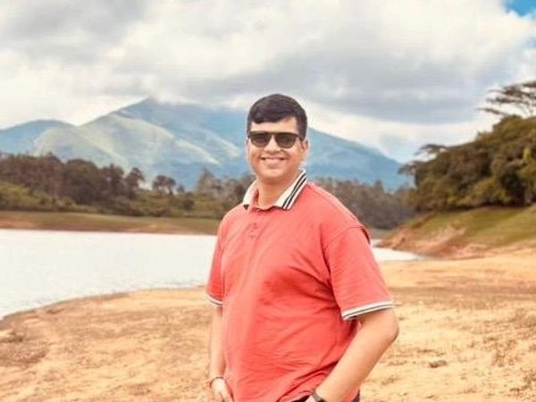 Man in sunglasses standing by a lakeside with mountains in the background.