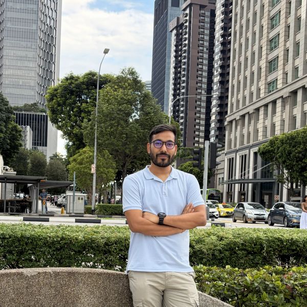 Man in light blue polo shirt standing outdoors in an urban area with tall buildings.