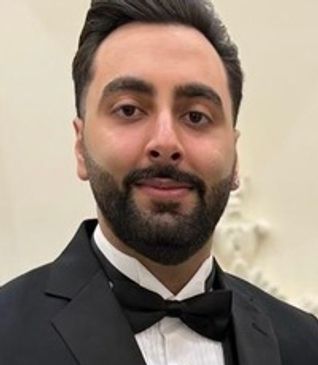 A young man with black hair and a beard wearing a suit, smiling slightly at the camera. 