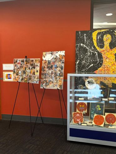 Art display with collage boards and a glass case featuring various artworks against an orange wall.