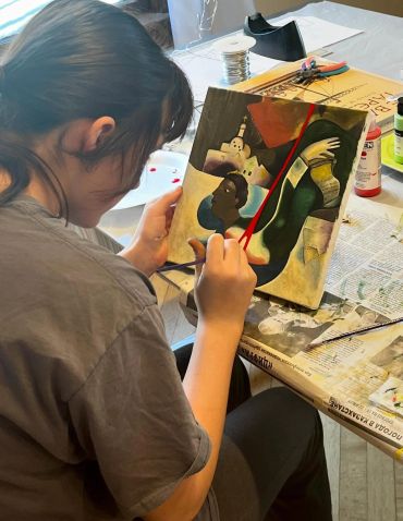 A person painting a colorful abstract artwork at a cluttered table.