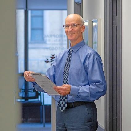 Smiling man in blue shirt and tie holding a clipboard in an office corridor.