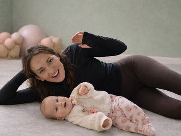 A smiling woman lying on the floor with a baby in cozy clothes.