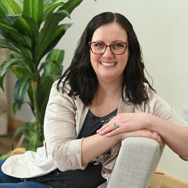 Smiling woman with glasses sitting on a chair indoors near a plant.