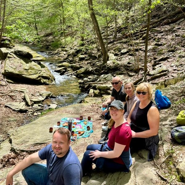 Group of people sitting beside a cascading creek in the woods with some snacks nearby on a rock.