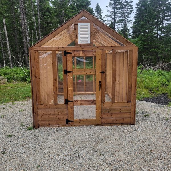 Small wooden greenhouse with clear plastic panels in a forest setting.