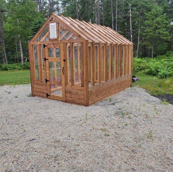 A wooden greenhouse with transparent panels in a forest clearing.