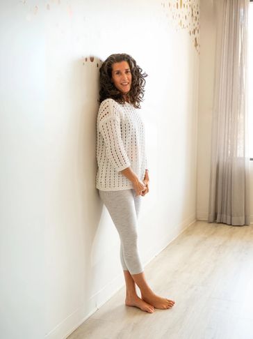 Holly is leaning against the wall, dressed in a white sweater and grey and white leggings. 