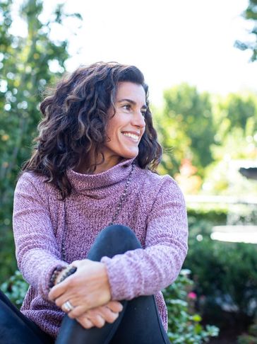 Holly is dressed in a purple sweater and black leggings, sitting outdoors, holding her knee. 