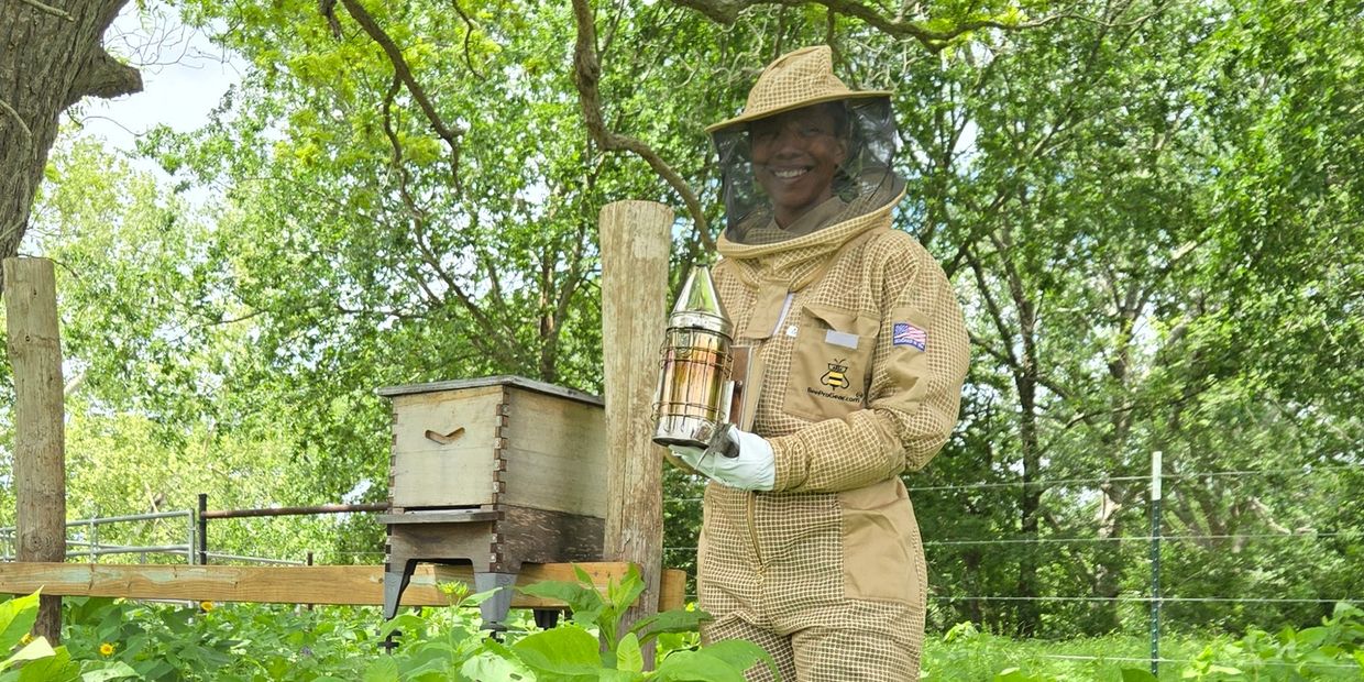 Smiling beekeeper in protective suit holding a smoker near a beehive outdoors.