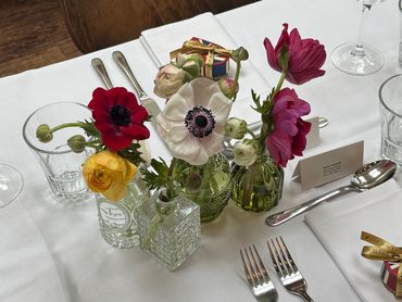 Colorful flowers in small glass vases arranged on a white tablecloth with dining utensils.