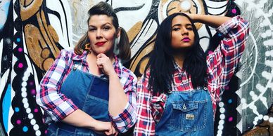 Two women posing in denim overalls and plaid shirts against a graffiti wall.
