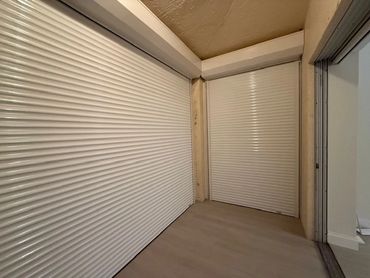 Two white rolling shutters in a corner space with beige flooring.