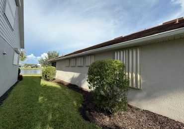 Side of a house with hurricane shutters and trimmed bushes under a blue sky.
