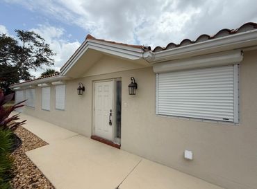House exterior with closed white hurricane shutters and beige walls under a cloudy sky.