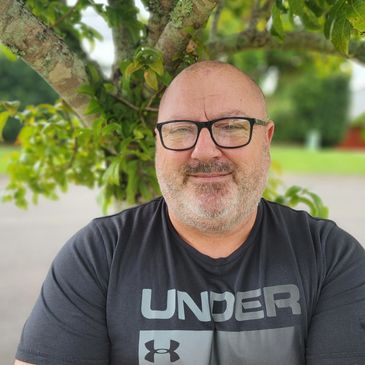 Headshot of John next to tree with arms folded. John's wearing glasses, has stubble and a wry smile