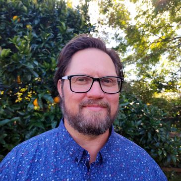 Headshot of Luke outdoors. Luke wears glasses, has brown hair, a full beard and a bright blue shirt.