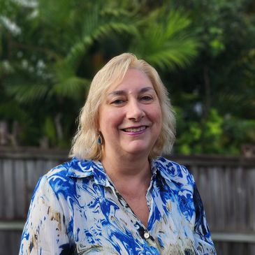 Headshot of Michelle standing near trees. Michelle is blonde, smiling broadly and is wearing blue