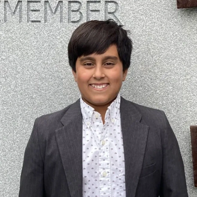 Smiling young person in a suit jacket standing against a gray wall.